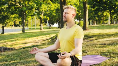 Lotus pozisyonunda yoga minderinde oturan genç adam dışarıda yoga ve meditasyon yapmaya hazırlanıyor. Fitness ve sağlıklı konsept.