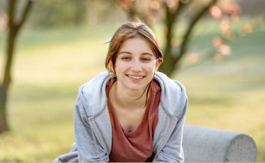 Bahar parkında oturan, gülümseyen ve kameraya bakan genç bir kızın portresi. Sıradan stil.