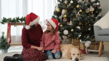 Anne ve kız Noel Baba şapkalı Golden Retriever köpeğinin yanında Noel ağacının altında oturuyor ve mutlu bir şekilde konuşuyor.