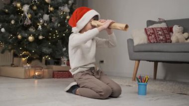 Kırmızı şapkalı küçük çocuk evde Noel ağacının altında otururken kağıttan dürbünlü dürbüne bakıyor.