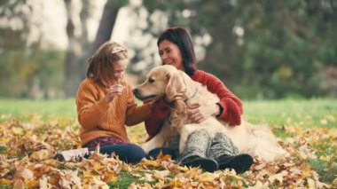 Anne ve kızı sonbahar parkında altın renkli köpekle eğleniyorlar. Mutlu aile dışarıda köpeklerle oynuyor..