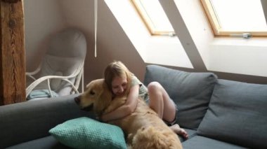 Evdeki kanepede uzanmış Golden Retriever köpeğine sarılan ve gülümseyen kız çocuğu. Sevimli çocuk, çatı katında safkan evcil köpek besliyor.