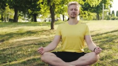Lotus pozisyonunda yoga minderinde oturan genç bir adam kapalı gözlerle dışarıda yoga yapmaya hazırlanıyor. Fitness ve sağlıklı konsept.