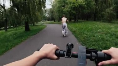 Paten kayan genç bir kızla parkta elektrikli scooter süren genç bir kadın. Modern araçlı arkadaşlar doğa aktivitelerinin keyfini çıkarıyorlar.
