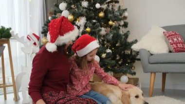 Küçük kızıyla birlikte Golden Retriever köpeğinin yanında oturmuş, süslenmiş Noel ağacının altında...