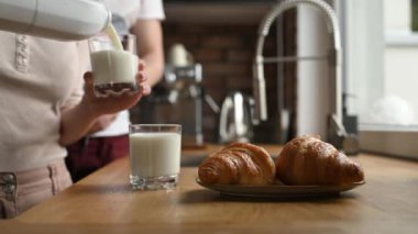Bardağa süt döken ve mutfakta kruvasanla kahvaltı yapan bir kız. Beyaz organik latte ve Fransız pastası olan kadın ve erkek çift.