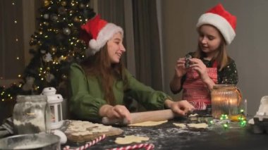 Şenlikli şapkalı mutlu kızlar zencefilli kurabiyelere hamur hazırlarken süslü odada noel ağacıyla merdane kullanıyorlar.