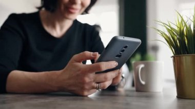 Genç bir kadın internette akıllı telefon kullanıyor ve mutfakta gülümsüyor.