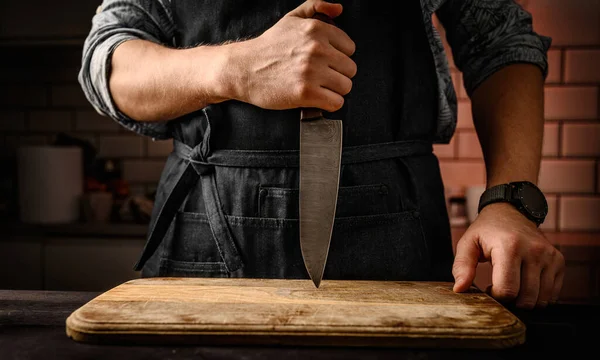 Chief man in apron stubs knife blade to wooden board at kitchen. Restaurant professional with culinary tool