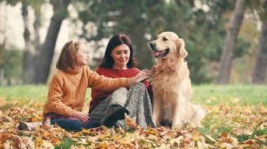 Anne ve kızı sonbahar parkında eğleniyor ve altın renkli köpekleri okşuyor. Mutlu aile dışarıda köpeklerle oynuyor..