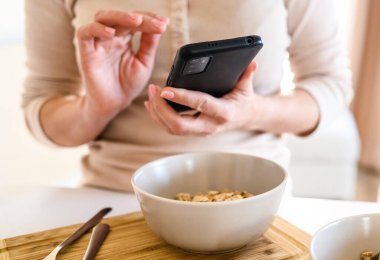 Girl with oatmeal bowl and smartphone in hands checking social media news during cereal breakfast. Woman with mobile phone and granola at kitchen