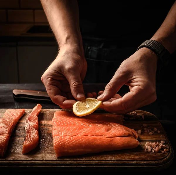 キッチンで木製テーブルにレモンスライスでサーモンフィレットを準備するチーフハンド 健康的な夕食のために赤い魚ステーキを調理する男 — ストック写真