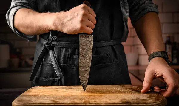 Chief man in apron holding knife and stubs blade to wooden board at kitchen. Restaurant proffesional with culinary tool