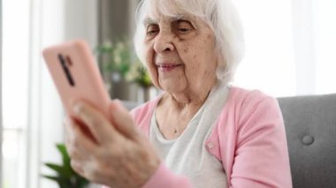 Yaşlı kadın çocuklarla video aracılığıyla konuşuyor. Parlak odada akıllı telefon kullanıyor.