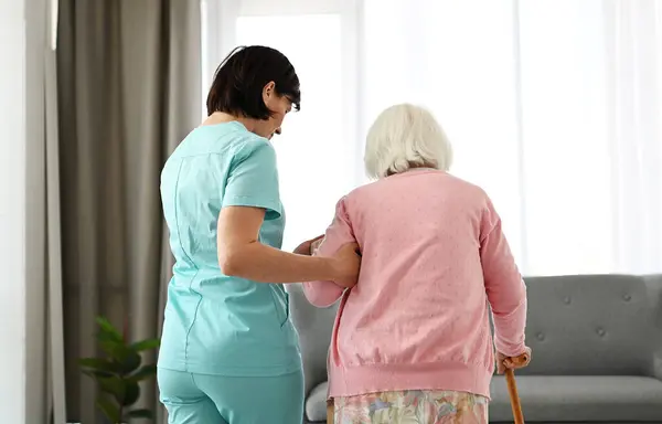Nurse Helps Elderly Woman Move Around Room, Embodying Concept Of Care And Support For Old People
