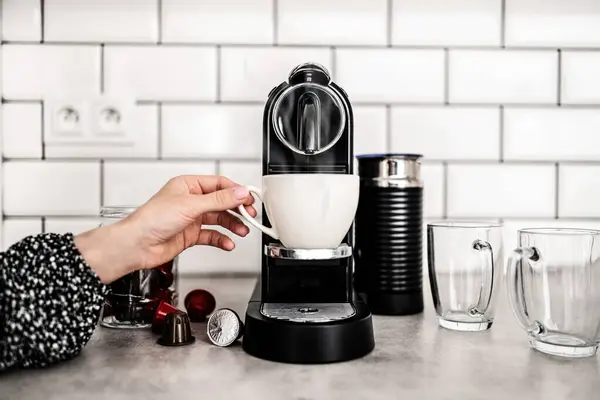 Girl hand with capsule coffee machine and white cup at kitchen. Woman preparing italian caffeine beverage using professional espresso maker