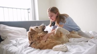 Neşeli küçük kız, tatlı köpeğiyle yatakta oynuyordu, ikisi de mutlu bir şekilde gülümsüyordu.
