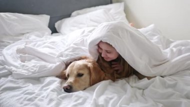 Yatakta battaniyenin altında tatlı bir köpekle eğlenen neşeli genç kız.