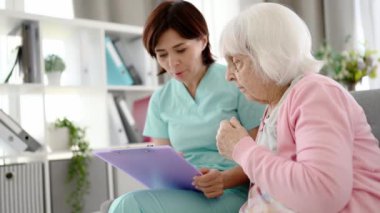 Sağlık işçisi, aydınlık bir odadaki yaşlı bir kadına ait sağlık belgelerini okuyor.