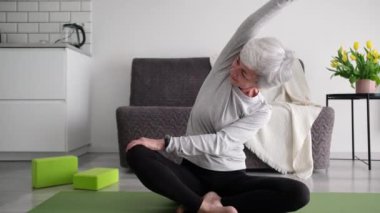 Gri saçlı, spor yapan ve evde yoga yapan yaşlı bir kadın.