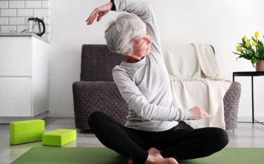 Gri saçlı, spor yapan ve evde yoga yapan yaşlı bir kadın.