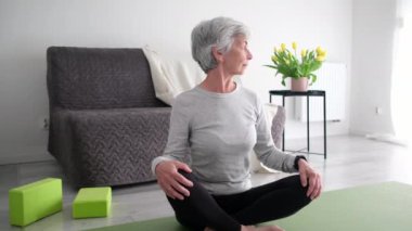 Gri saçlı yaşlı kadın evinde yoga minderi üzerinde boynu esnetiyor.