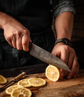 Şef eller, mutfaktaki tahtada bıçakla limonu dilimlere ayırıyor, yakın çekim manzarası var. Limonata hazırlığı için turunçgil meyvesi