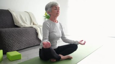 Enerjik yaşlı kadın evde yoga yapıyor, Lotus pozisyonunda oturuyor.