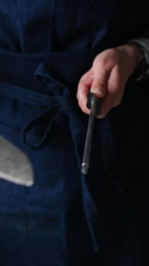 Patron önlüklü kız büyük bir bıçağı keskinleştiriyor bir çubuk aletle, dikey görünüm