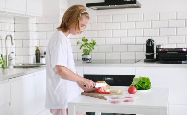 Küçük bir kız mutfakta bıçakla tahtada domates kesiyor.