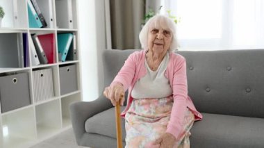 Oturma odasındaki koltukta oturan yaşlı kadın. Cane gülüyor ve kameraya bakıyor.