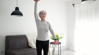 Gri saçlı, çekici ve aktif yaşlı bir kadın evde dambıllarla spor yapıyor.