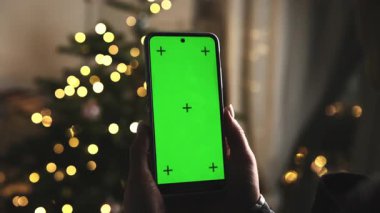 Kızın elinde Noel ağacı ışıklarına karşı yeşil ekranlı akıllı bir telefon var.