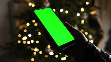 Kızın elinde Noel ağacı ışıklarına karşı yeşil ekranlı akıllı bir telefon var.