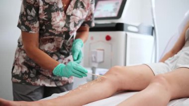 Doktor bir kozmetoloji kliniğinde lazer saç çıkarma prosedürü sırasında kızların bacaklarına jel sürüyor.