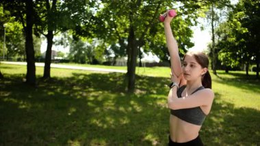 Parkta spor yapan ve sabah rutininin bir parçası olarak dambılları kaldıran formda bir kız.