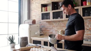 Genç adam, kahvaltıya hazırlanmak için kasede yumurta sallarken mutfakta omlet pişiriyor.