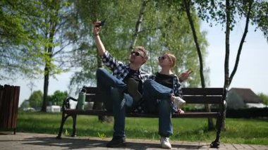 Baba ve kızı bir bankta oturup yaz parkında selfie çekiyorlar.