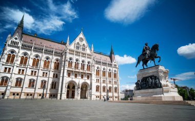 Hungarian Parliament Building In Budapest With A Statue Against Clouds In The Sky