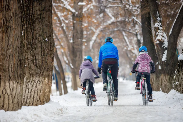 Father and children ride bicycles in winter. A man with his son and daughter ride bicycles along the bike path in the winter park. View from the back