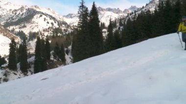 A bearded man with a backpack descends a snowy slope in the mountains. A tourist with a backpack and trekking poles walks along the trail
