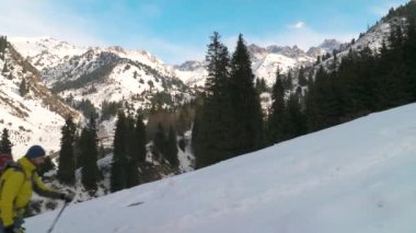 A bearded man with a backpack is actively climbing the trail in winter in the mountains. Hiker with backpack and trekking poles walking outdoors in winter mountains