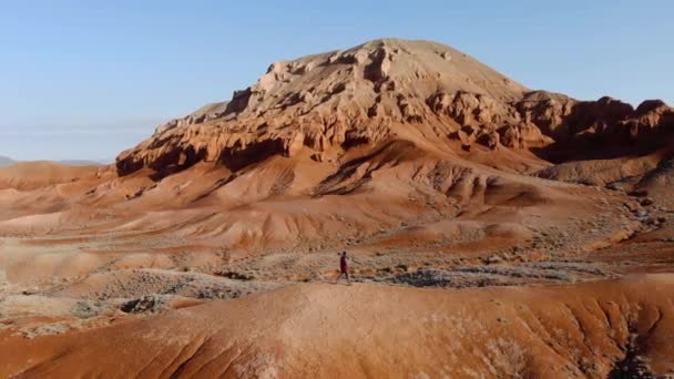 写真家は珍しい赤い岩の山々を背景に丘の上を早朝に歩きます 三脚の上にカメラを持った男が赤いダウンジャケットを着ていた カザフスタンのAzhirzhar Tract — ストック動画
