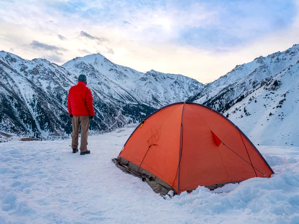A male tourist stands early in the morning in the snowy mountains near the red tent in which he spent the night. A traveler in a red down jacket travels high in the mountains. View from the back