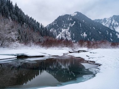 Dağlı kış manzarası ve yansımalı donmuş bir nehir. Bulutlu karlı bir gün. Yumuşak ışıklandırma