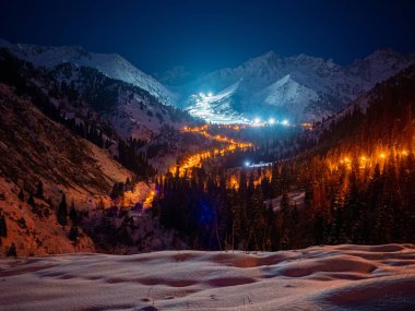 Aydınlatılmış yılan yolunun ve Chimbulak kayak merkezinin gece panoraması. Kış akşamının sonlarında dağların tepesinde