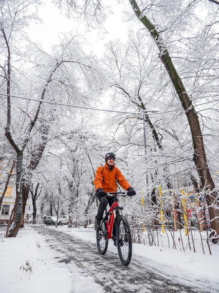 A man rides a bicycle through a winter city among snow covered trees. The guy on the red bike is wearing an orange jacket and a bicycle helmet on his head. Active lifestyle in winter