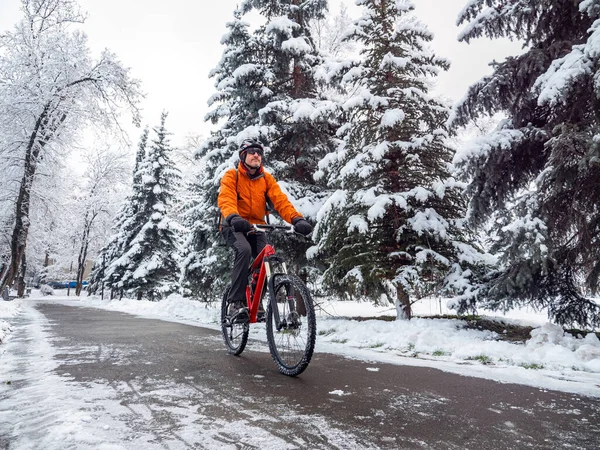 A guy rides a bicycle along the road among snow covered trees. A man on a red bicycle with a bicycle helmet on his head. Active lifestyle in winter