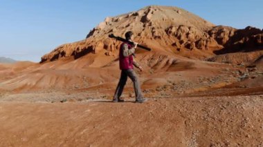 Fotoğrafçı sabahın erken saatlerinde bir tepenin üzerinde sıra dışı kızıl kayalık dağların arkasında yürür. Kırmızı ceketli, tripodunda kamera olan bir adam. Azhirzhar Tract, Kazakistan