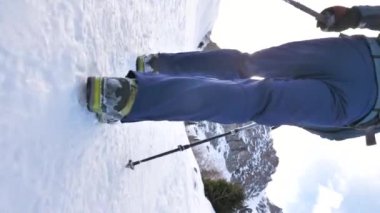 Rüzgarlı bir kış gününde dağlarda yürüyen bir adamın dikey video görüntüleri. Kar kırıntıları yerde uçuşuyor. Sırt çantalı ve sırıklı bir adam.